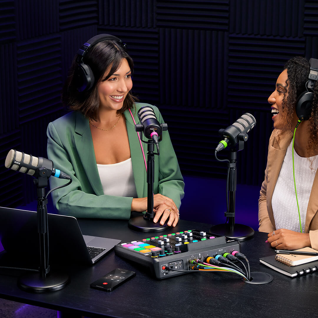 Two podcast hosts talking into microphones with a RØDECaster Pro II and a laptop on the desk.