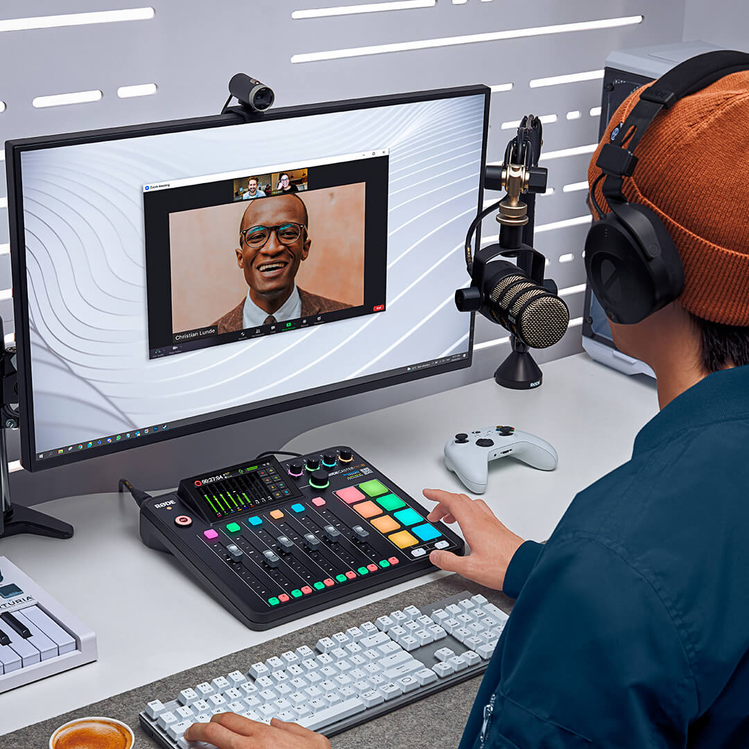 Streamer at a desk using a RØDECaster Pro II, with a video call on the monitor, a PodMic on a boom arm and a game controller beside it.